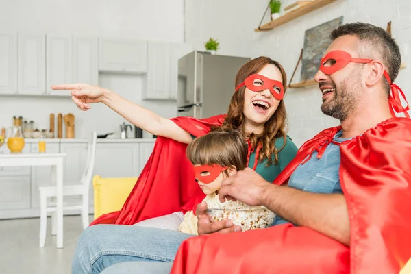 Fröhliche Mutter zeigt mit dem Finger, warum Familie in Kostümen von Superhelden auf dem Sofa sitzt und fernsieht — Stockfoto