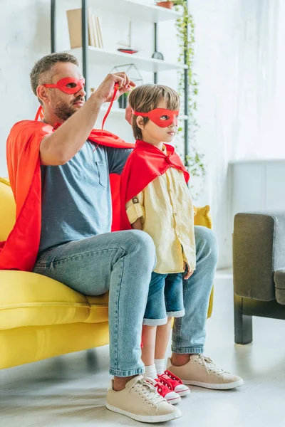 Отец в костюме супергероя надевает красную маску на сына, сидя дома на диване — стоковое фото