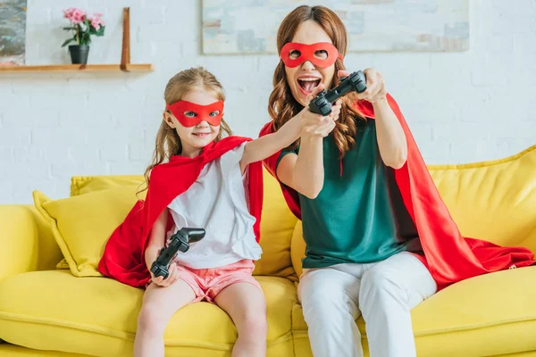KYIV, UKRAINE - JULY 5, 2019: Excited woman and cute child in costumes of superheroes playing video game while sitting on couch at home — Stock Photo