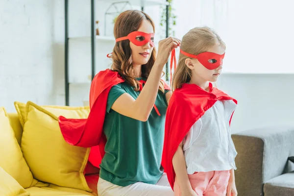 Sonriente mujer poniendo traje de superhéroe en adorable hija - foto de stock