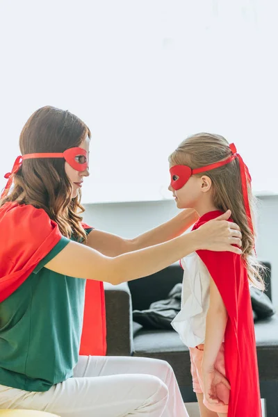 Mãe e filha em trajes de super-heróis olhando um para o outro em casa — Fotografia de Stock