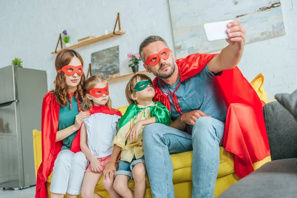 Familia en trajes de superhéroes tomando selfie en el teléfono inteligente mientras se sienta en el sofá en casa - foto de stock