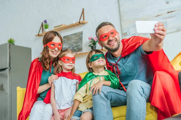 Happy family in costumes of superheroes taking selfie on smartphone while sitting on couch at home — Stock Photo