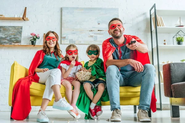 Glückliche Familie in Kostümen von Superhelden vor dem Fernseher, während sie auf dem Sofa mit Popcorn sitzt — Stockfoto