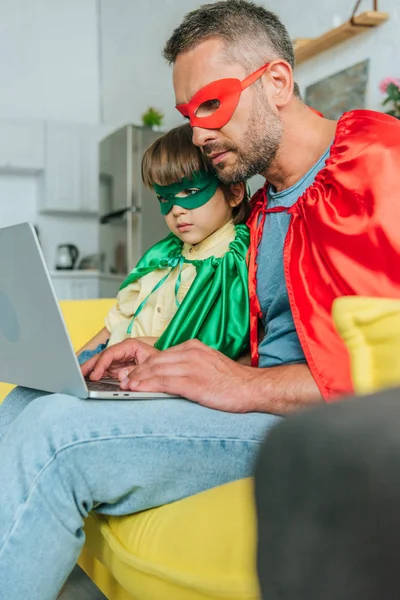 Selective focus of attentive father and son in superheroes costumes using laptop at home — Stock Photo