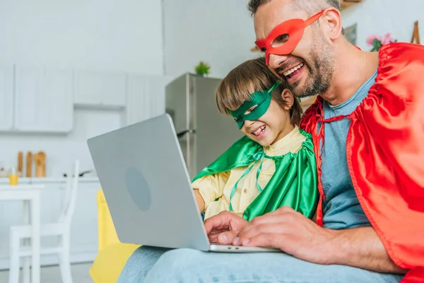 Веселый отец и сын в костюмах супергероев с ноутбуком дома — стоковое фото