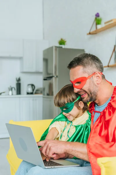 Sonriente padre e hijo en trajes de superhéroes usando el ordenador portátil en casa - foto de stock