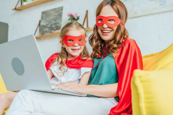 Alegre mamá y su hija en trajes de superhéroes mirando a la cámara mientras se utiliza el ordenador portátil en casa - foto de stock