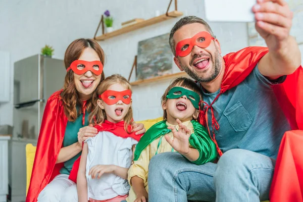 Selective focus of happy family in superheroes costumes sticking tongues out while taking selfie on smartphone at home — Stock Photo