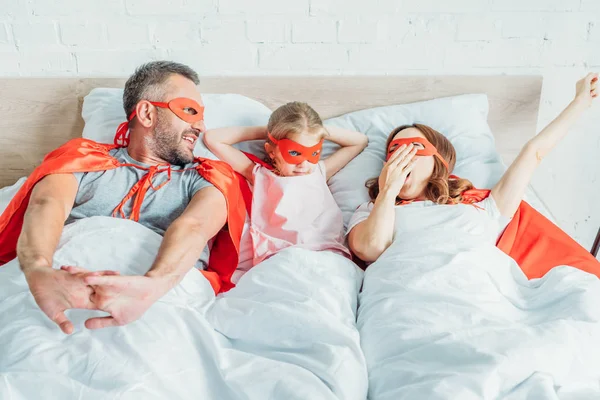 Familia feliz bostezando y estirándose mientras está acostado en la cama en trajes de superhéroes - foto de stock