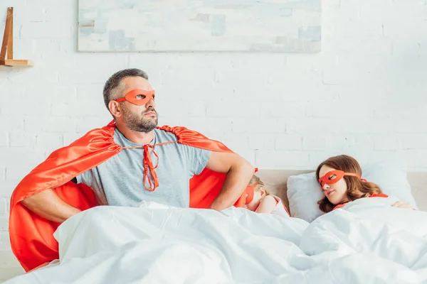 Pai sério no traje de super-herói sentado na cama perto da família dormindo, de mãos dadas nos quadris e olhando para longe — Fotografia de Stock