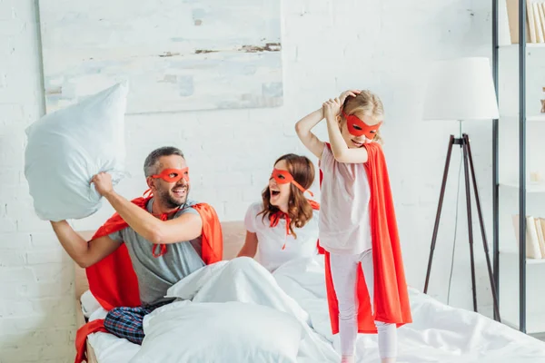 Alegre padre disfrazado de superhéroe sosteniendo almohada mientras está sentado en la cama cerca de la esposa y la hija - foto de stock