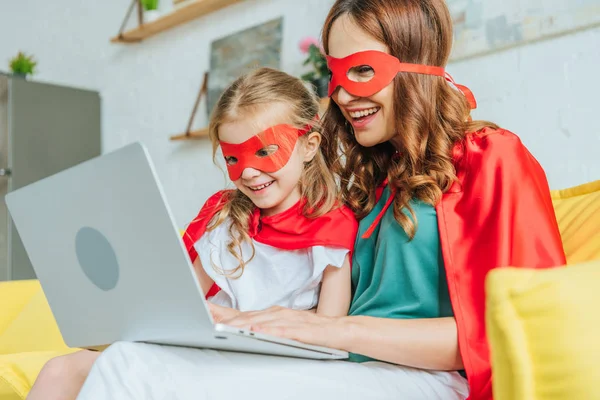 Щаслива жінка з дочкою в костюмах супергероїв, використовуючи ноутбук вдома — стокове фото