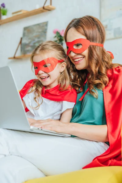 Alegre mamá y su hija en trajes de superhéroes utilizando el ordenador portátil en casa - foto de stock