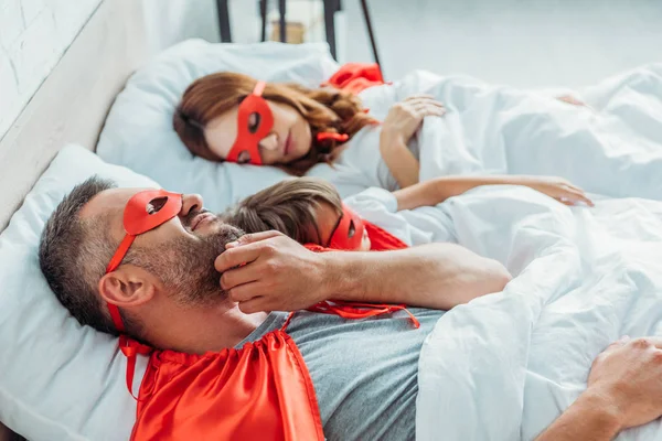 Foco seletivo do homem em traje de super-herói coçando bochecha enquanto deitado na cama perto de esposa e filho — Fotografia de Stock