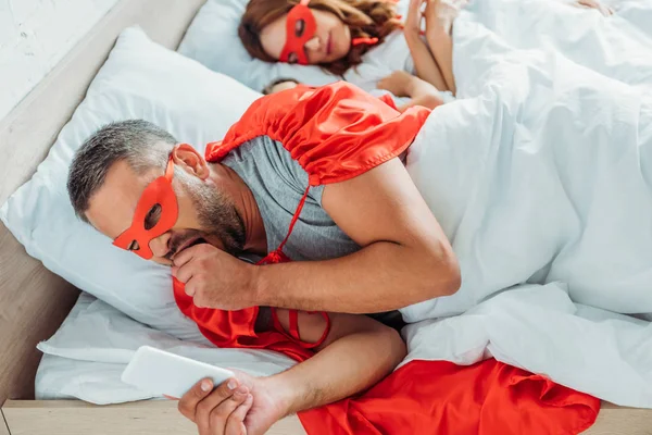 Foco seletivo do homem em traje de super-herói bocejando enquanto deitado na cama perto da família adormecida — Fotografia de Stock