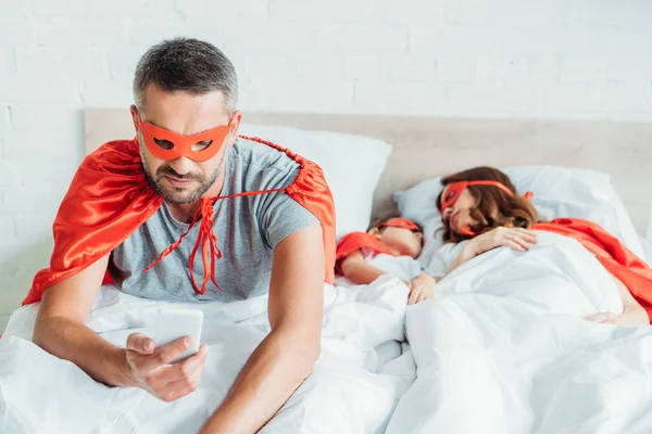 Man in costume of superhero using smartphone while sitting in bed near sleeping family — Stock Photo