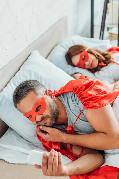 Messa a fuoco selettiva dell'uomo in costume di supereroe utilizzando smartphone mentre si trova a letto vicino moglie e figlio — Foto stock