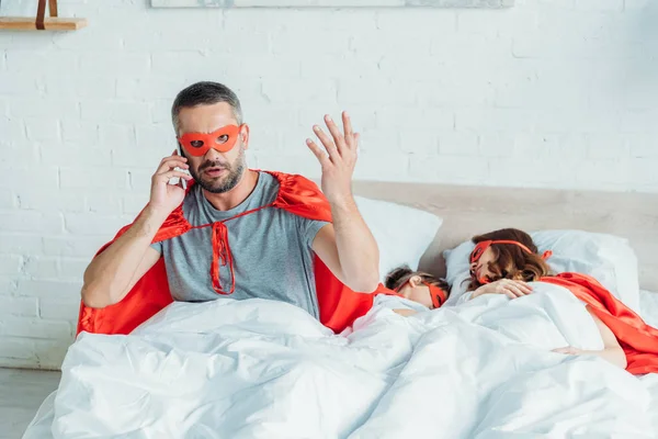 Homme en costume de super-héros parler sur smartphone et gestuelle tout en étant assis dans le lit près de la famille endormie — Photo de stock