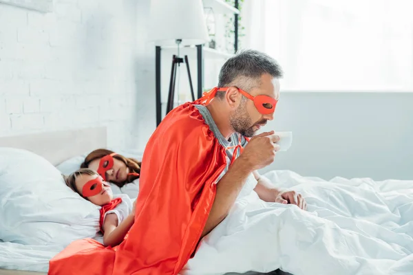 Father in costume of superhero drinking coffee while sitting in bed near sleeping wife and son — Stock Photo