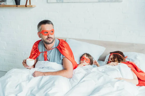 Мужчина в костюме супергероя пьет кофе и смотрит в сторону, сидя в постели рядом со спящей женой и сыном — стоковое фото