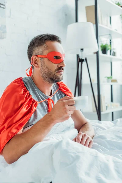 Hombre pensativo disfrazado de superhéroe bebiendo café y mirando hacia otro lado mientras está sentado en la cama - foto de stock