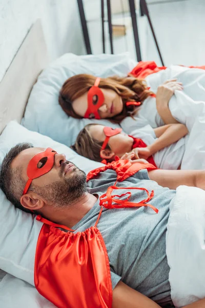 Selective focus of mom, dad and son sleeping in bed in superheroes costumes — Stock Photo