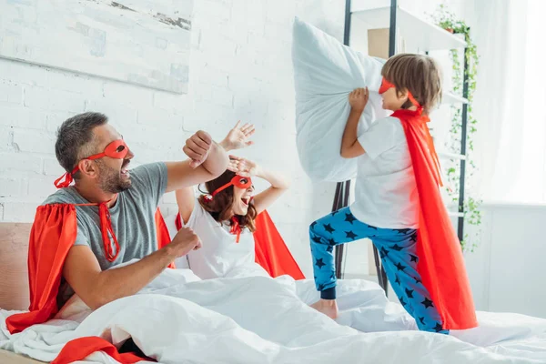 Padres alegres en trajes de superhéroes luchando con el hijo sosteniendo la almohada - foto de stock