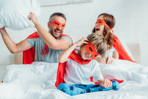 Счастливая семья в костюмах супергероев, дерущихся с подушкой в постели — стоковое фото