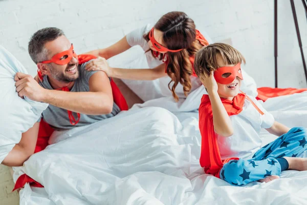 Familia alegre en trajes de superhéroes luchando con almohada en la cama - foto de stock