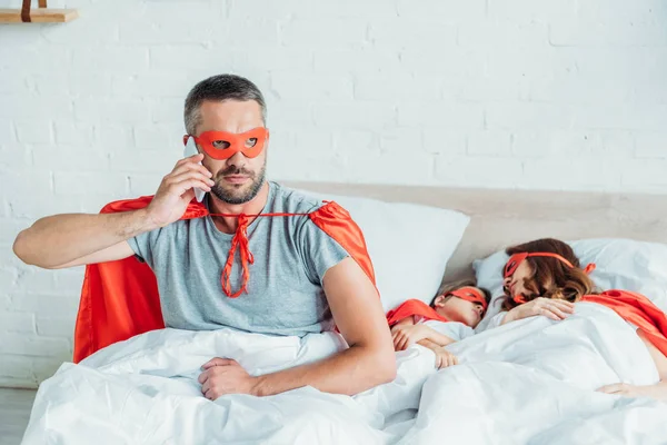 Serious man in costume of superhero talking on smartphone while sitting in bed near sleeping wife and son — Stock Photo