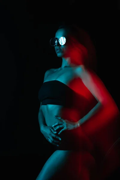 Movimento sfocatura di donna attraente in occhiali da sole in piedi con mano sul fianco su nero — Stock Photo