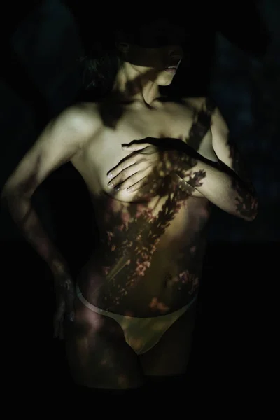 Обрізаний вид молодої жінки, що покриває груди, стоячи з рукою на стегні на чорному з тінями — стокове фото