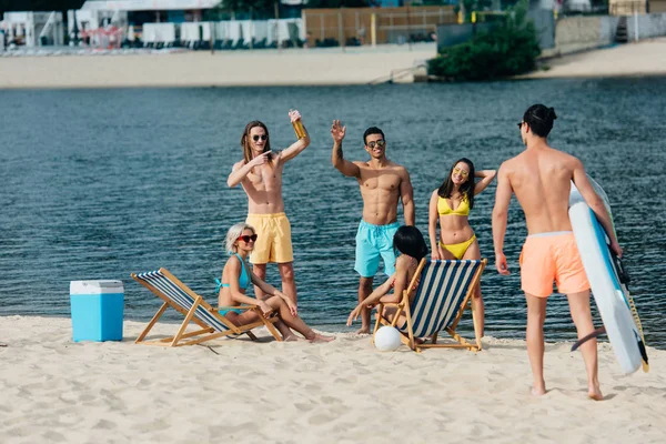 Весёлые мультикультурные друзья машут руками спортсмену, держащему доску для серфинга на пляже — стоковое фото