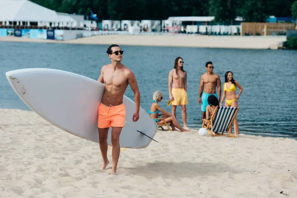 Bel homme tenant la planche de surf tandis que des amis multiculturels reposant sur le bord de la rivière — Photo de stock