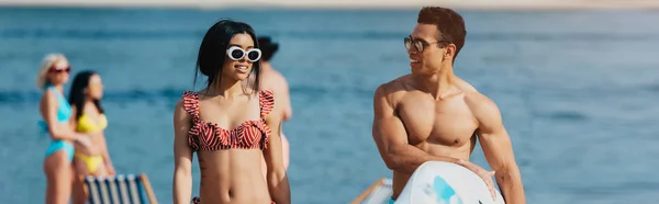 Panoramic shot of handsome man holding surfboard near attractive woman in swimsuit — Stock Photo