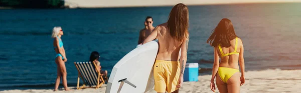 Back view of young man holding surfboard near girl in swimsuit, panoramic shot — Stock Photo