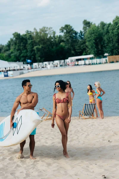 Fröhlich Mixed Race Mann hält Surfbrett in der Nähe afrikanisch-amerikanisches Mädchen im Badeanzug — Stockfoto