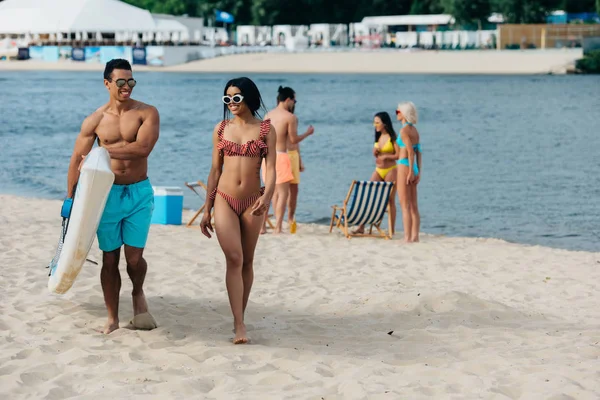 Handsome mixed race man holding surf board while walking near african american girl in swimsuit — Stock Photo