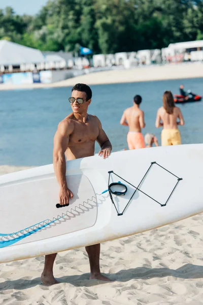 Handsome mixed race man holding surfing board and looking away on beach — Stock Photo