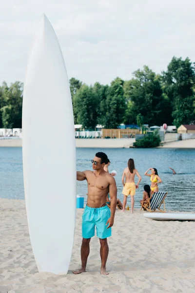 Bonito misto raça homem de pé perto surf bordo na praia — Fotografia de Stock