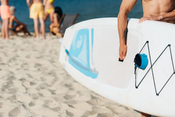 Cropped view of young man holding surfing board while standing on beach — Stock Photo