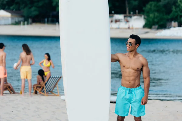 Uomo di razza mista sorridente e bello in piedi vicino al surf sulla spiaggia — Foto stock