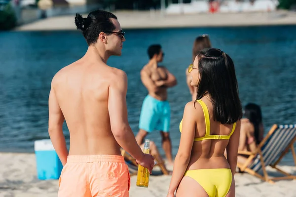 Back view of young man with bottle of beer standing near girl in swimsuit on beach — Stock Photo