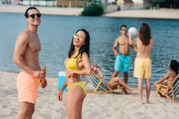 Alegre joven hombre y mujer con botellas de cerveza sonriendo a la cámara mientras está de pie en la playa cerca de amigos multiculturales - foto de stock