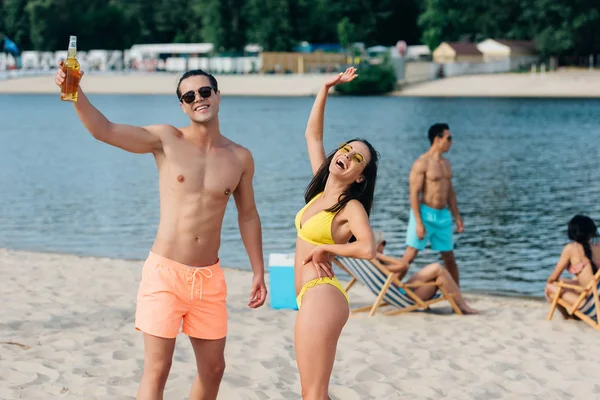 Attraktive Mädchen lächelnd und winkende Hand, während sie am Strand neben einem fröhlichen Mann mit einer Flasche Bier steht — Stockfoto