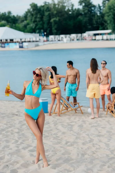 Fröhliche, attraktive Frau im Badeanzug, die eine Flasche Bier hält, während sie neben multikulturellen Freunden steht — Stockfoto