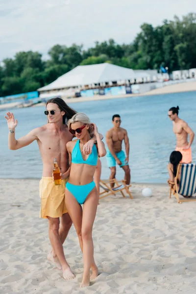 Joven guapo saludando de la mano y abrazando a la novia mientras está de pie en la playa cerca de amigos multiculturales - foto de stock