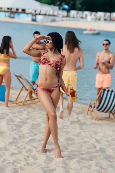 Beautiful african american woman in swimsuit looking away while standing on beach near multicultural friends — Stock Photo