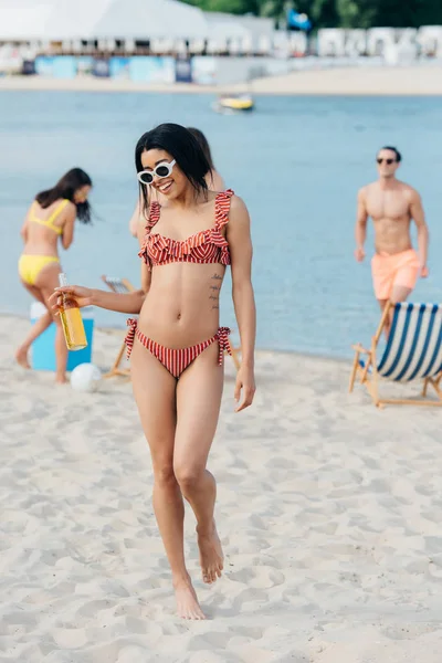 Attraktive Afroamerikanerin im Badeanzug, die am Strand steht und eine Flasche Bier in der Hand hält — Stockfoto